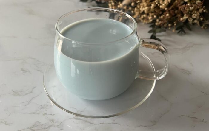 A glass cup and saucer on a marble table top, containing a pale-blue coloured latte, displayed with dried lavender flowers out of focus in the background