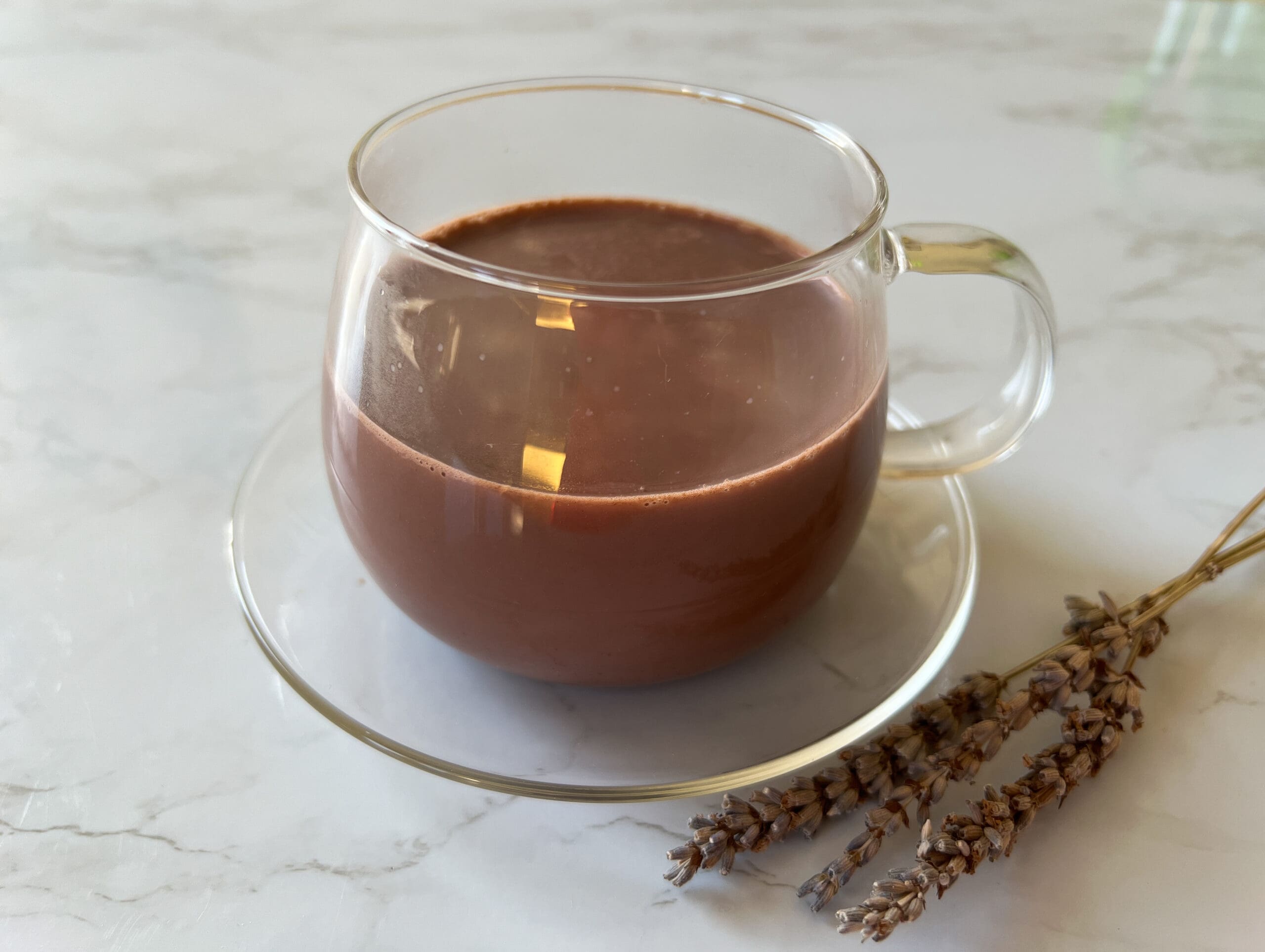 Hot chocolate served in a glass cup and saucer displayed on a marble table top with sprigs of dried lavender next to it