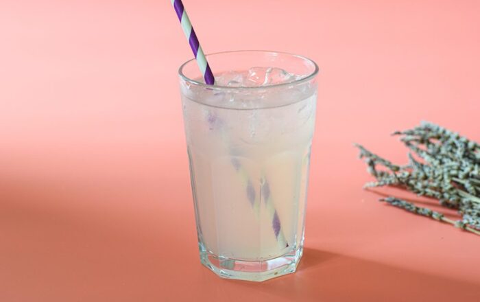 A glass of lavender lemonade on a peach coloured background with a red and white paper straw