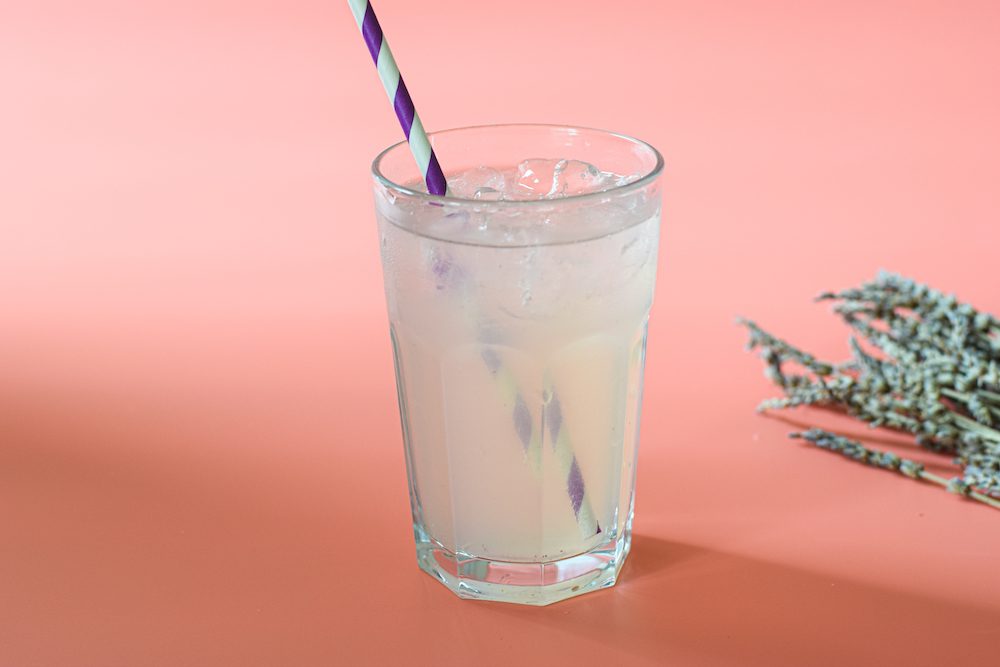 A glass of lavender lemonade on a peach coloured background with a red and white paper straw