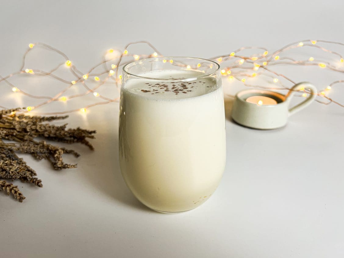 A glass of lavender eggnog displayed on a pale off-white table with a pine branch, a sugar bowl and a tangle of gold fairy lights