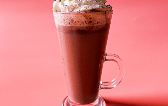 A tall glass mug of lavender hot chocolate topped with whipped cream displayed on a pink background