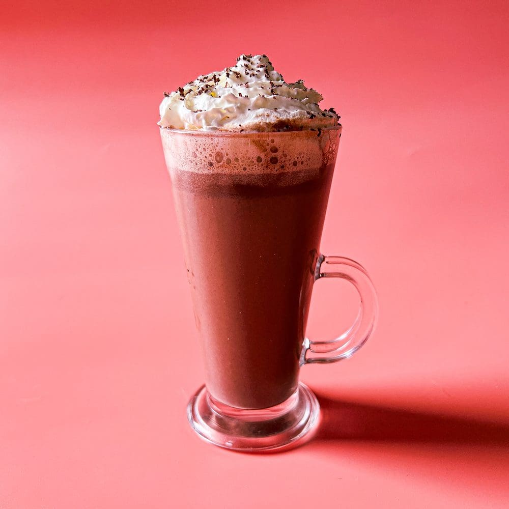 A tall glass mug of lavender hot chocolate topped with whipped cream displayed on a pink background