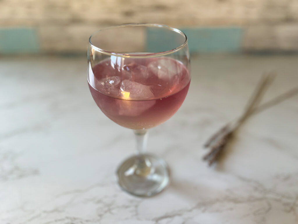 A pink cocktail in a stemmed glass with ice, on a marble table top, with dried lavender in the background